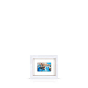 A framed artwork featuring a London docklands scene, with a blue sky and warehouses, float-mounted in a white frame.